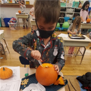 Kindergartner with mask on digging out inside of pumpkin as his desk 